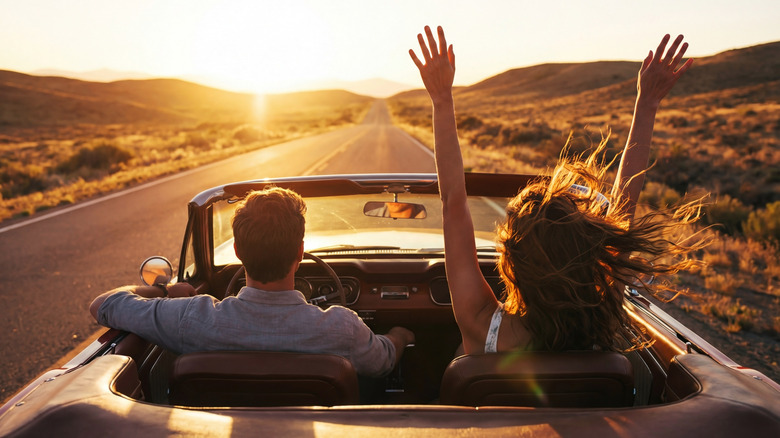 Young couple driving convertible through desert road at sunset with her arms in the air