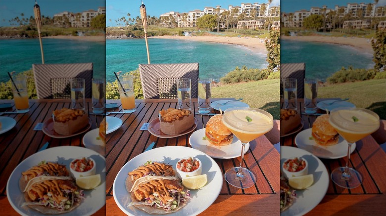 Dishes and drinks served on a wooden table overlooking the water at Merriman's Kapalua
