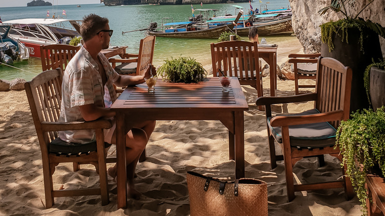 A man sits at a table on a beach under a rock overhang looking at boats in the water