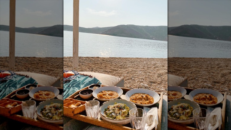 A wooden tray holds multiple dishes and glasses on the sand near sparkling water