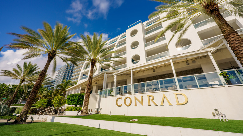 View the Fort Lauderdale Beach Conrad Hotel as seen from the street, with gold logo and palm trees on a sunny day