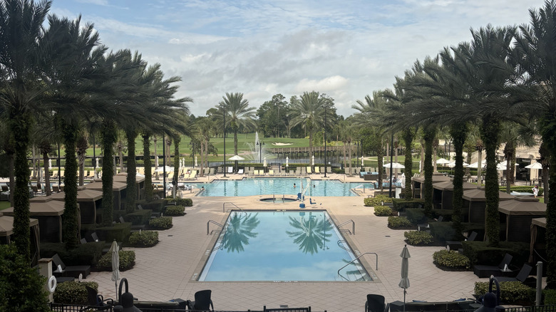 View of the elegant pool area at the Waldorf Astoria Orlando with palm trees and cabanas lining either side, and the property's golf course in the background