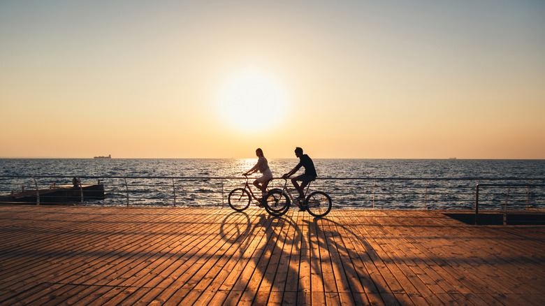 Two people bicycle on a wooden walkway with a large body of water in the background during sunset