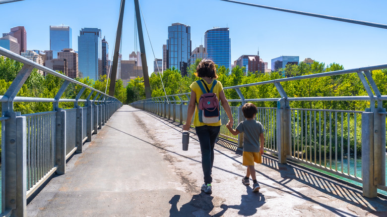 Mother and son are walking on a pedestrian bridge in calgary holding hands, enjoying the view of the bow river and the skyscrapers of the downtown