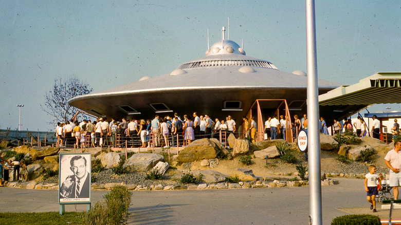 View of visitors waiting on line outside the Braniff Airways Space Ship flying saucer attraction in the Satellite City exhibit at the Freedomland USA theme park, located in the Baychester section of The Bronx, New York City, September, 1961. A sign visible in the lower left foreground advertises upcoming Harry James Orchestra performances at the Moon Bowl from September 29 to October 1, 1961.