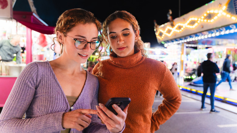 Two young adult women viewing content on a mobile phone during a fun night out at a theme park