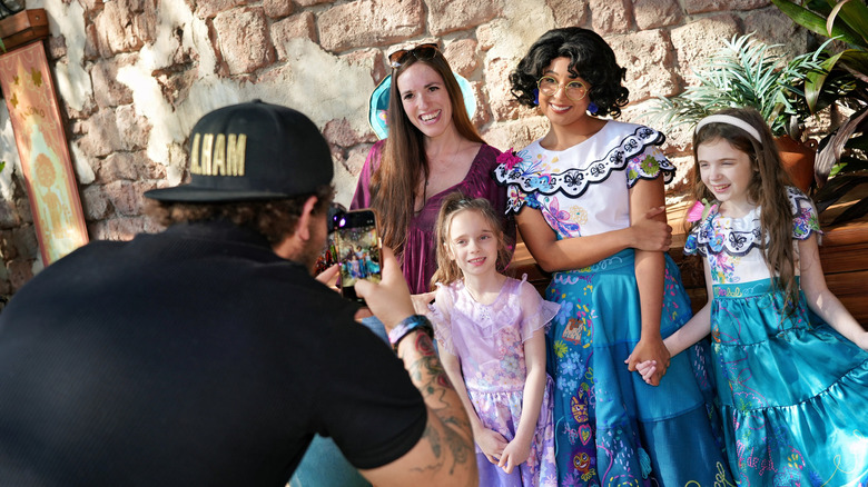 Mom and two daughters pose with Mirabel at Magic Kingdom while dad takes a photo on a phone