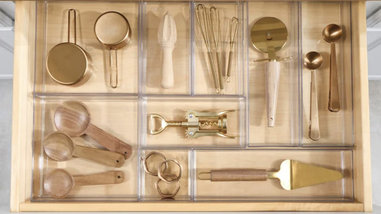 View of pulled out light wood drawer featuring modular acrylic drawer organizers to store gold and wood-tone kitchen utensils