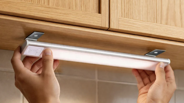 Close-up view of hand installing undercabinet light bar on light wood cabinetry