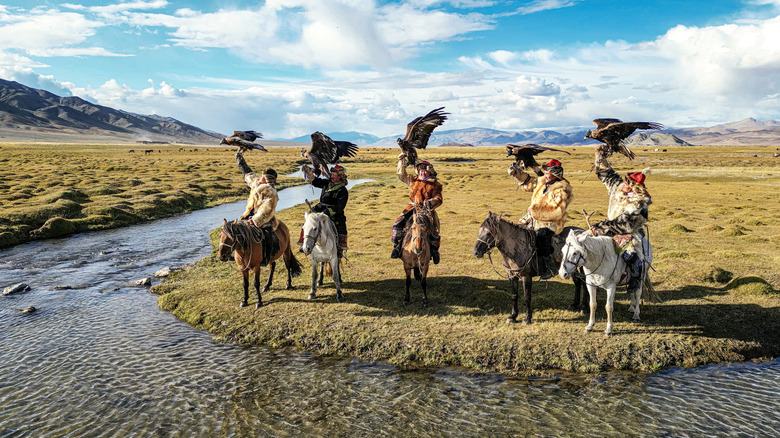 Group of nomadic Mongolian eagle hunters on horseback with golden eagles spreading wings in front of river Bayan Olgii, West Mongolia