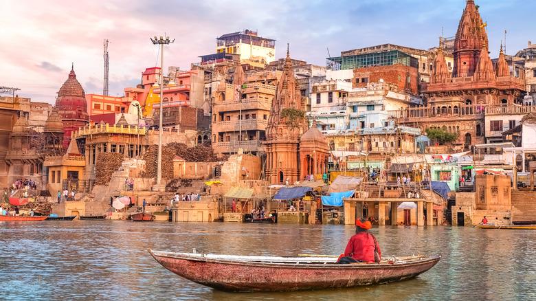 Ancient Varanasi city architecture and Ganges river ghat at sunset with view of an Indian sadhu sitting a boat.