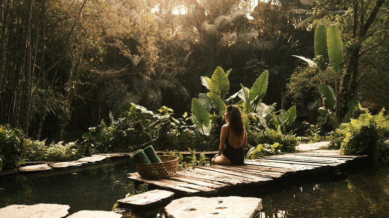 Woman sitting on a wooden pier by a natural pond, in Indonesia finding tranquility and wellness in a lush tropical resort