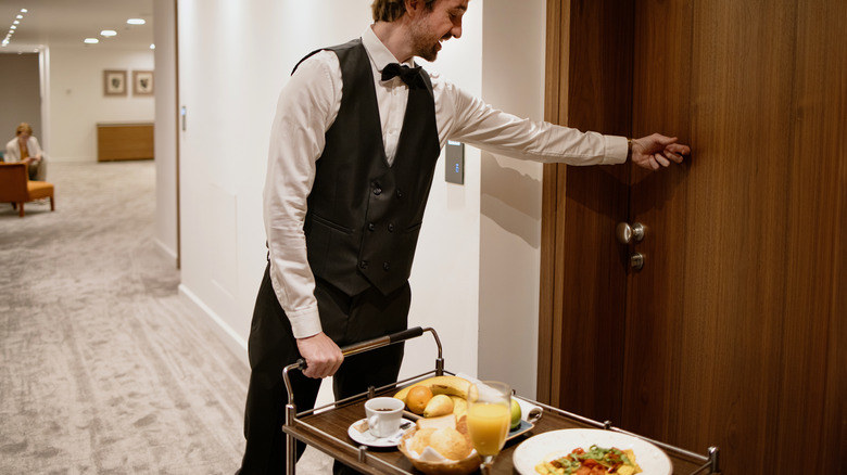 Waiter in uniform delivering tray with food in a room.