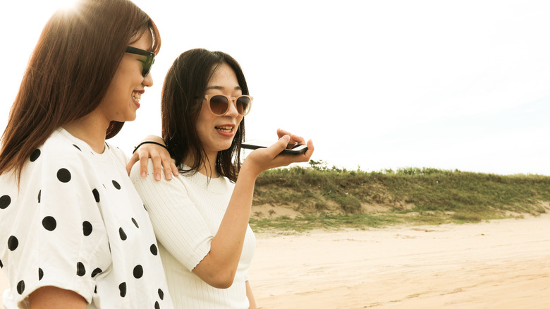 Two women on a beach, one of them is holding a smartphone and speaking into it