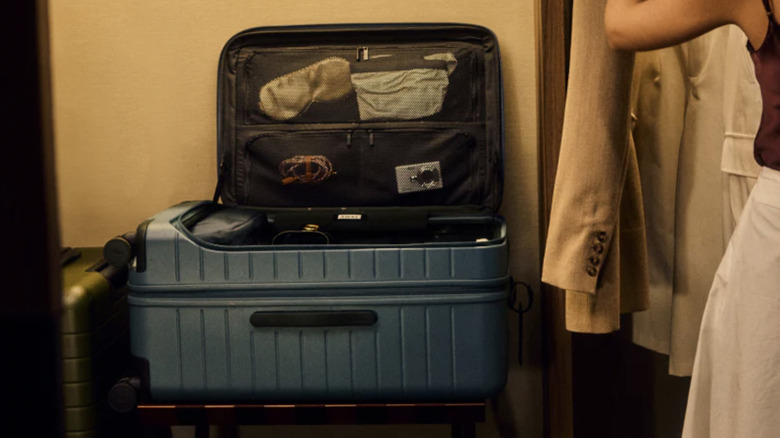 Close up view of open Away brand suitcase on a luggage holder while a woman hangs clothing in a hotel closet