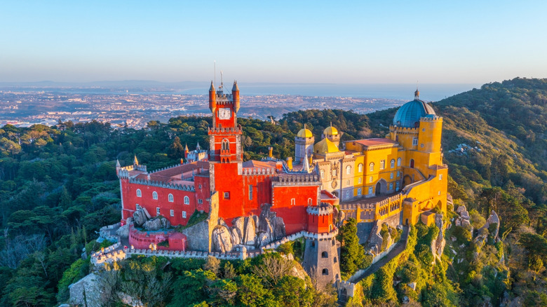 Bright red castle, and a yellow palace, tower over lush green hills, sea in far horizon
