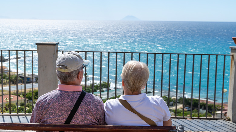 Two elderly people sit on a bench overlooking the sea in Calabria, with clear skies and the Aeolian Islands visible in the distance