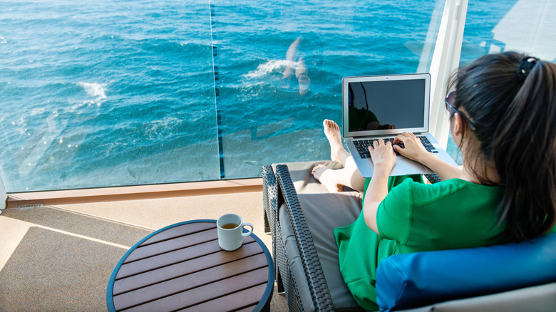 Back view of Asian woman with ponytail working on a laptop on her cruise ship balcony with bright blue water in the background