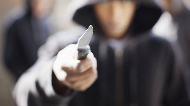 Blurred man in a black hoodie menacingly holding an in-focus locking blade knife towards the camera
