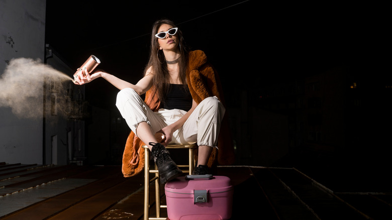 Fashionable young woman sits on a chair with her feet on a suitcase, spraying hairspray into the air