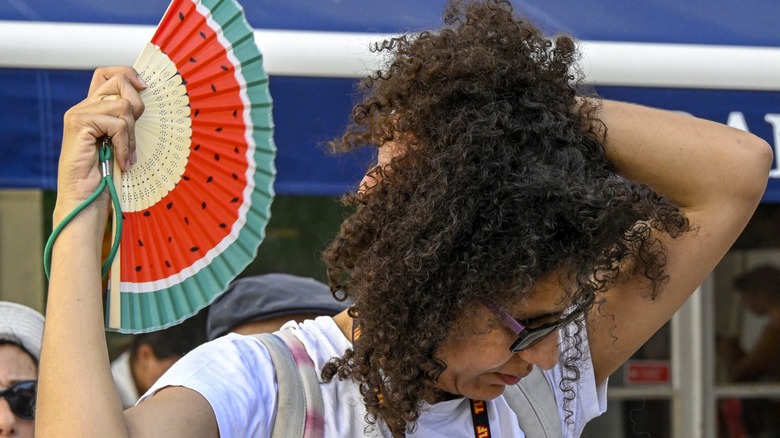 A woman fans the back of her neck with a watermelon-colored accordion fan