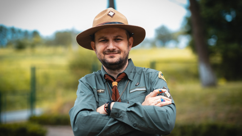 A park ranger in uniform is standing with arms crossed, smiling in an outdoor setting