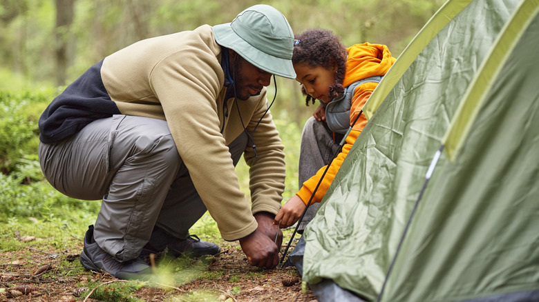 A Black man and child set up a tent together in forest, crouching and working as team