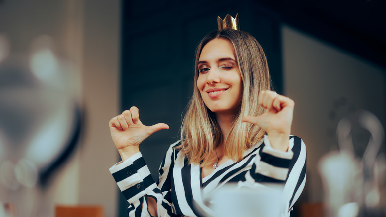 Cheerful woman with a gold crown on her head pointing at herself with both thumbs