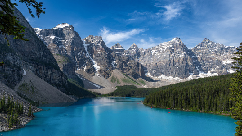 The turquoise waters of a lake in the foreground with the rugged peaks of the Canadian Rockies and dense evergreen forests in the distance