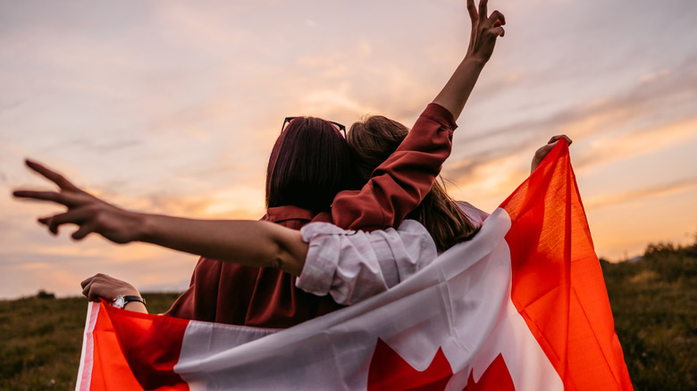 Two, young, females, covering themselves with Canadian flag. Standing on the meadow at sunset