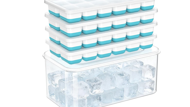 View of four silicon and plastic ice cube trays with lids stacked on top of plastic ice cube dispenser on white background