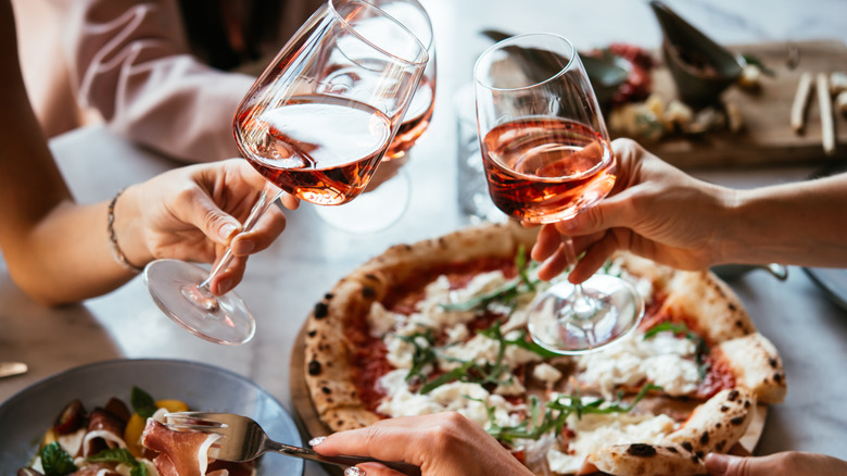 Two hands clinking glasses of wine over a pizza in the background