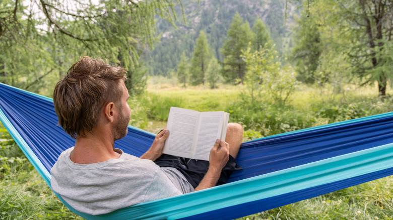A man reclines in a hammock while reading a book, surrounded by forest