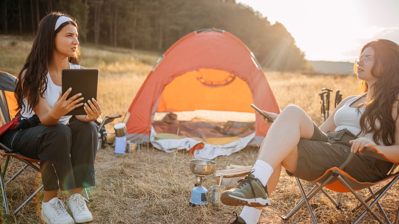Two young women sit outside of their tent, one holding a tablet as they discuss something