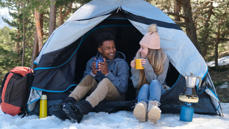 A cute couple sipping out of mugs by their tent surrounded by snow