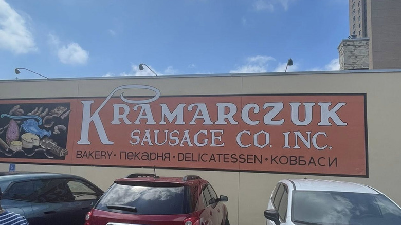 Exterior sign at Kramarczuk's Sausage with cars parked in front of it and blue sky in the background