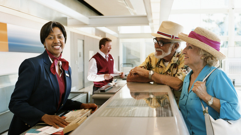 An elderly couple standing at a desk in front of a travel agent, smiling at the camera
