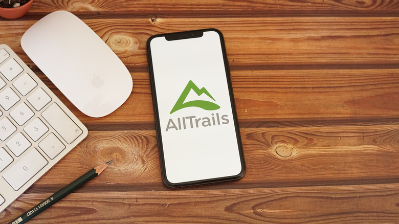 A close-up of the AllTrails logo on a phone screen next to a computer on a wooden table