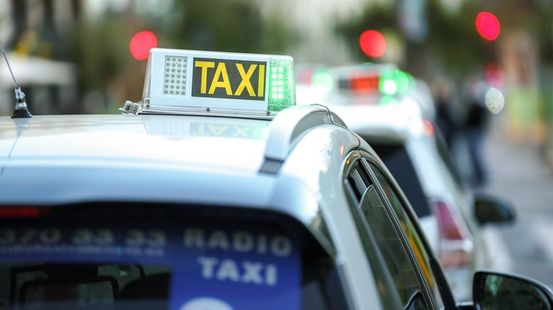 The top of a white taxi with a yellow taxi sign moving through traffic with one window open