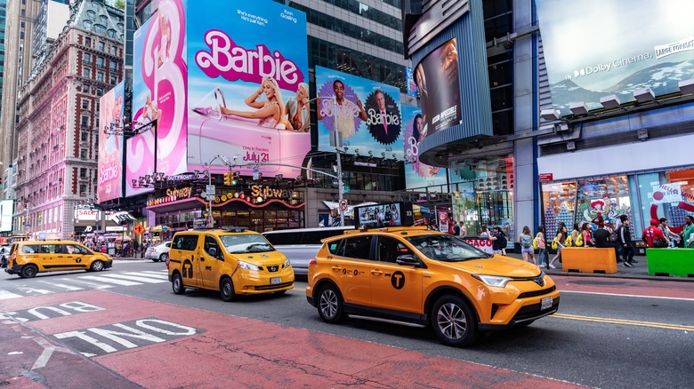 Yellow taxis driving through Times Square, NYC, with billboards for a "Barbie" movie in the background