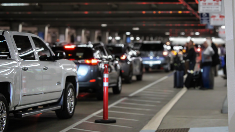 Cars lined up in front of an airport entrance, with people waiting with suitcases in the background