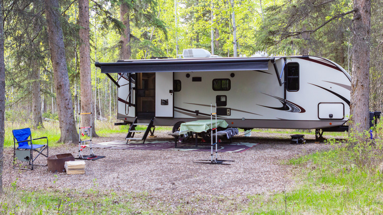 A campsite all set up in a wooded area with a large travel trailer, campfire, chair, and picnic table