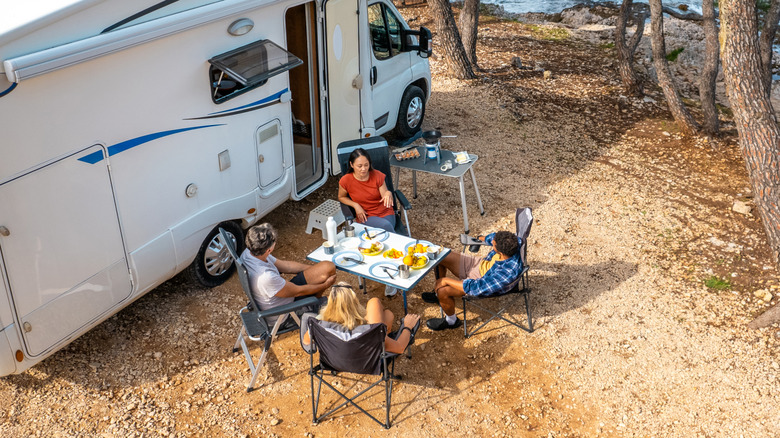 Diverse group relaxes at coastal campsite, sharing meals beside a parked RV