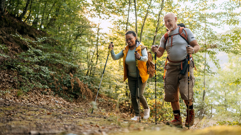 A smiling older couple hikes along a forest trail with trekking poles and backpacks