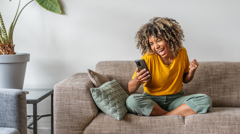 Excited happy young black woman holding smart phone device sitting on sofa at home