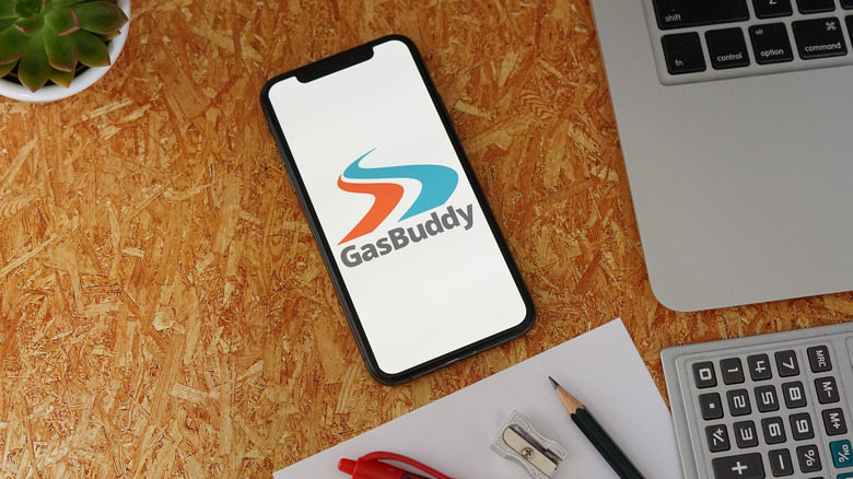 A phone and other items sit on a wooden surface with the GasBuddy app on the screen