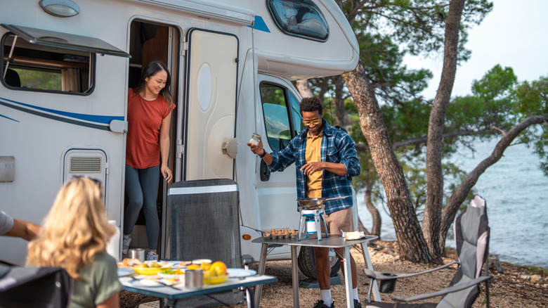 A group of friends gather outside of a motorhome to eat breakfast near a lakeshore