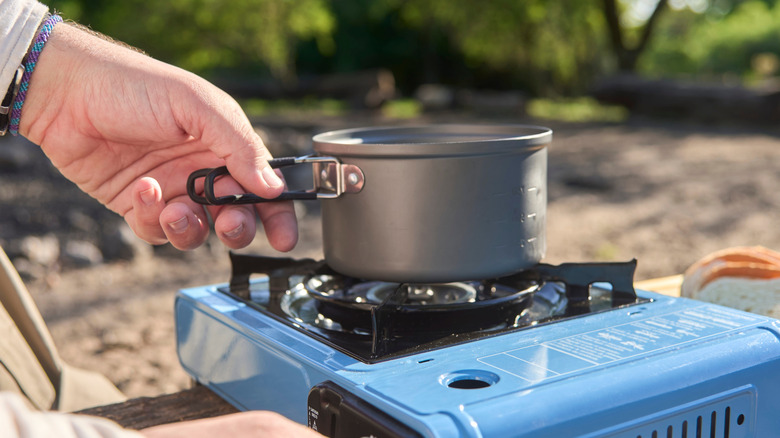 traveler cooking outdoors while camping at a nature campground a sunny morning. Close up view, one hand lights the blue portable stove,