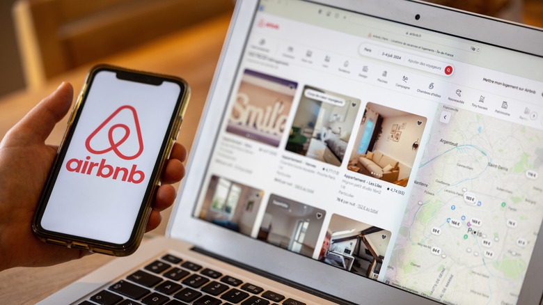 An individual holding a phone with a laptop open on Airbnb's website in the background