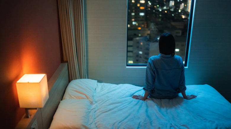 A shot of from behind of a woman sitting on a hotel bed looking out the window at night, with a hotel lamp open by the headboard
